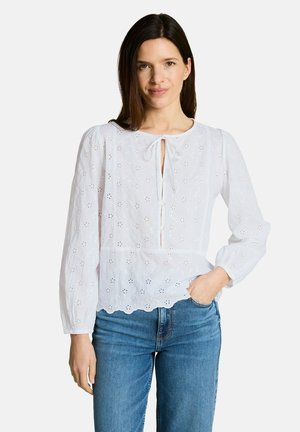 Woman with straight brown hair wears a white long-sleeve blouse with eyelet details and blue jeans, standing with one hand in pocket.