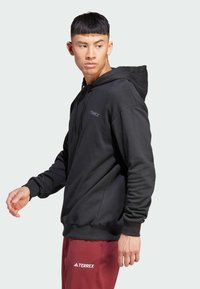 Sudadera con capucha negra de tela suave, con un bolsillo tipo canguro en la parte delantera y puños de canalé, combinada con pantalones burdeos.