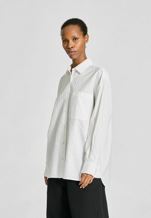 Femme aux cheveux courts portant une chemise blanche ample boutonnée et un pantalon noir, debout devant un fond clair uni.