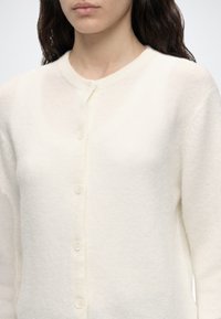 Femme portant un cardigan blanc doux à boutons avec des manches longues et un col rond sur un fond uni.