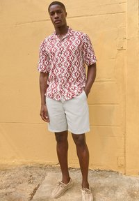 Short-sleeve shirt with a red and white floral pattern, paired with light gray shorts and beige woven shoes, standing against a yellow wall.