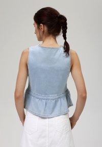 Lyseblå denim ermeløs topp med peplumkant, med rund hals og svakt frayed kanter. Bakutsikt viser en jevn tekstur.