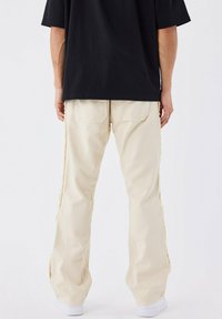 Pantaloni in denim beige con una vestibilità rilassata, caratterizzati da cuciture laterali sfrangiate e due tasche posteriori. Abbinati a una t-shirt nera a maniche corte.