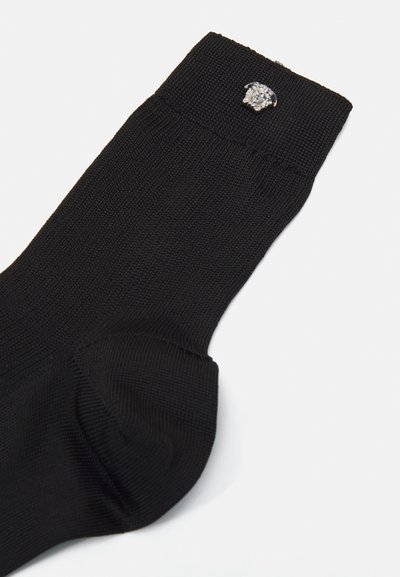 Čierna ponožka vyrobená z textúrovaného materiálu s rebrovaným lemom. Obsahuje kovový emblém na vrchu, ktorý zdôrazňuje minimalistické dizajnové detaily.