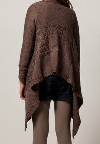 Brun stickad cardigan med intrikata vågmönster, öppen rygg och asymmetrisk nederkant. Texturerad tygkombination med åtsittande shorts och strumpbyxor.