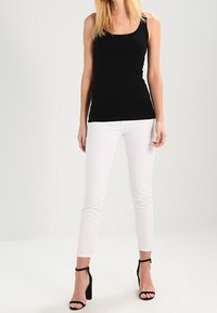 Débardeur noir, design ajusté, porté avec un jean slim blanc. Modèle associé à des escarpins noirs à bride cheville, se tenant devant un fond neutre.