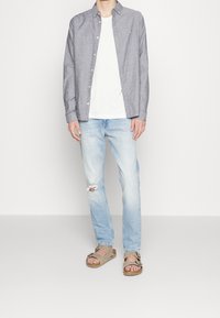 Men's outfit featuring a light gray button-up shirt over a white t-shirt, distressed light blue jeans, and beige sandals with a buckle.