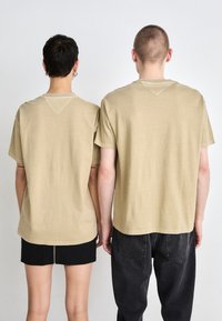 Beige puuvillatee -paidat rentoon istuvuuteen, joissa on lyhyet hihat ja pyöristetty kaula-aukko, nähtynä takaa. Mustat shortsit ja farkut ovat päällä.