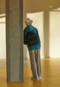 Rucsac cu model negru și gri purtat de o persoană îmbrăcată într-o hanorac teal și pantaloni bej, sprijinindu-se de un stâlp de beton într-un spațiu luminat de soare.