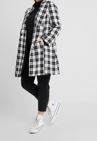 Abrigo de cuadros en blanco y negro con detalles en los bolsillos, diseño recto, material de mezcla de lana, usado sobre una camiseta y pantalones negros, combinado con zapatillas blancas.