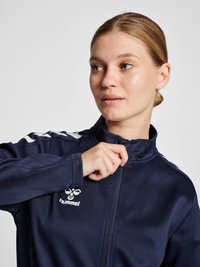 Marineblå zip-up sportsjakke med hvide striber på skuldrene, logo på brystet og glat stofstruktur; tætsiddende design med høj krave.