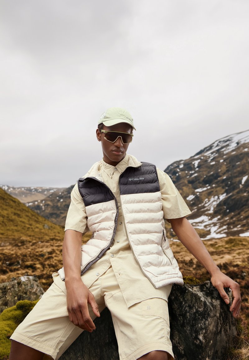 Gilet imbottito leggero beige e nero, maglietta beige a maniche corte e pantaloni corti beige. Gli accessori includono un cappellino chiaro e occhiali da sole.