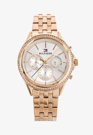 Ružovo zlaté kovové hodinky s puzdrom ozdobeným kryštálmi, bielym ciferníkom, tromi subciferníkmi a prepojeným remienkom. Zobrazuje logo Hilfiger.