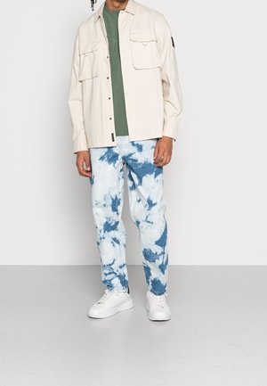 Hombre con sobrecamisa beige, camiseta verde, jeans teñidos anudados en azul y blanco, y zapatillas blancas de pie frente a un fondo blanco liso.