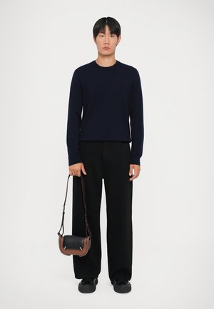 Suéter de manga larga azul marino, pantalones negros de pierna ancha y zapatos negros, sosteniendo un pequeño bolso cruzado con detalles en cuero marrón y negro.