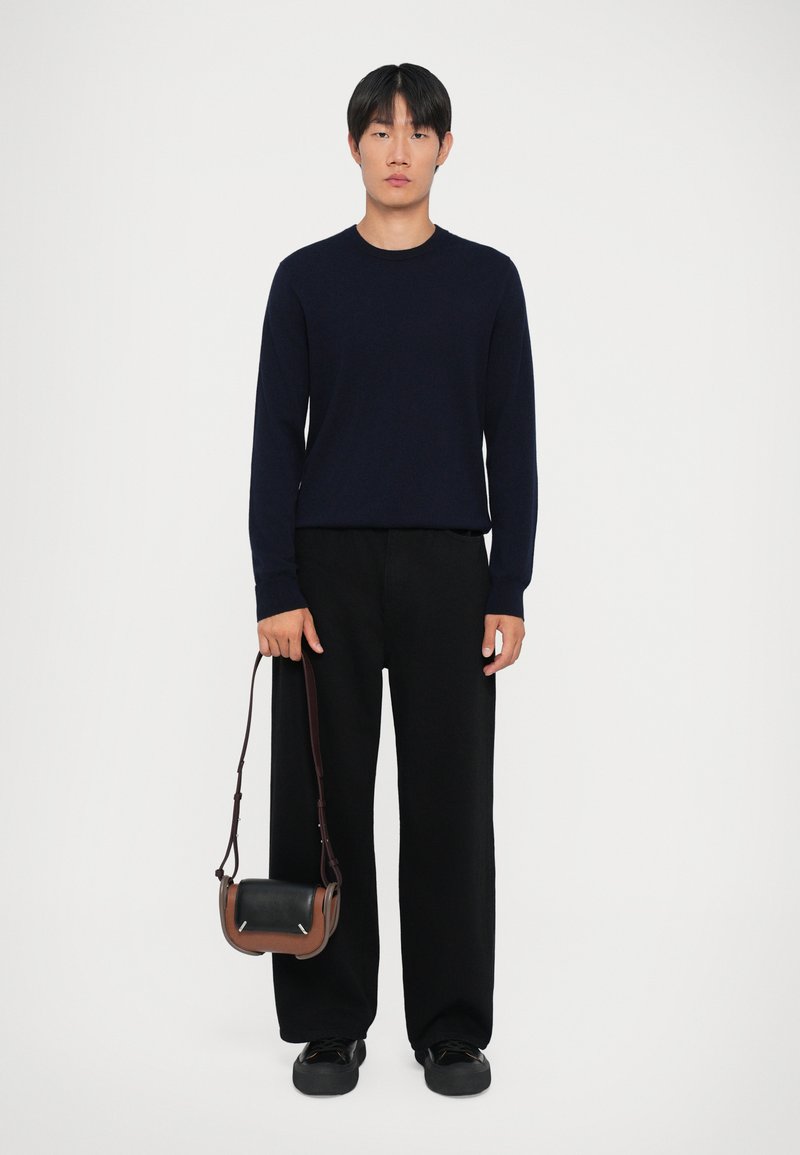 Maglione a maniche lunghe blu navy, pantaloni neri a gamba larga e scarpe nere, mentre tiene una piccola borsa a tracolla con dettagli in pelle marrone e nera.