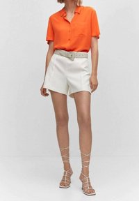 Pomarańczowa koszula z krótkim rękawem na guziki z kieszonką na piersi, zestawiona z białymi szortami z wysokim stanem i jasnymi sandałami na cienkich paseczkach.