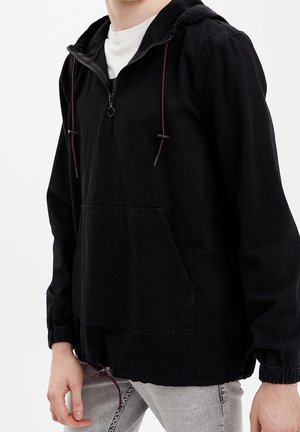 Osoba ubrana w czarną bluzę z kapturem z przednią kieszenią i czerwonymi sznurkami, w połączeniu z jasnoszarymi jeansami na jednolitym tle.