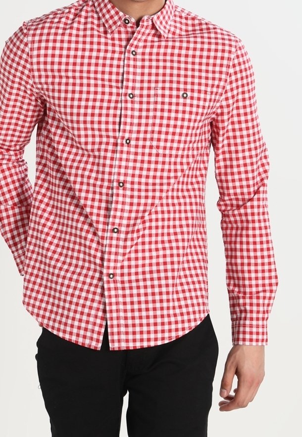 Homme portant une chemise à manches longues à carreaux rouges et blancs ainsi qu'un pantalon noir, debout devant un fond blanc uni.