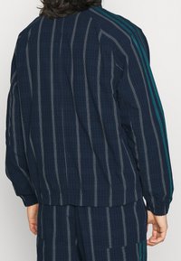 Mörkblå randig jacka med vertikala linjer och en texturerad tyg. Har teal ränder på axlarna samt elastiska manschetter och nederkant.