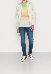 Mannenoutfit met een lichtgrijs overhemd met knopen, een multicolor gestreepte trui, blauwe jeans en bruine sneakers met witte zolen.
