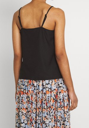 Vrouw draagt een zwarte mouwloze top en geplooide bloemenrok met blauwe, oranje en witte bloemen, staand met de rug naar een witte achtergrond.