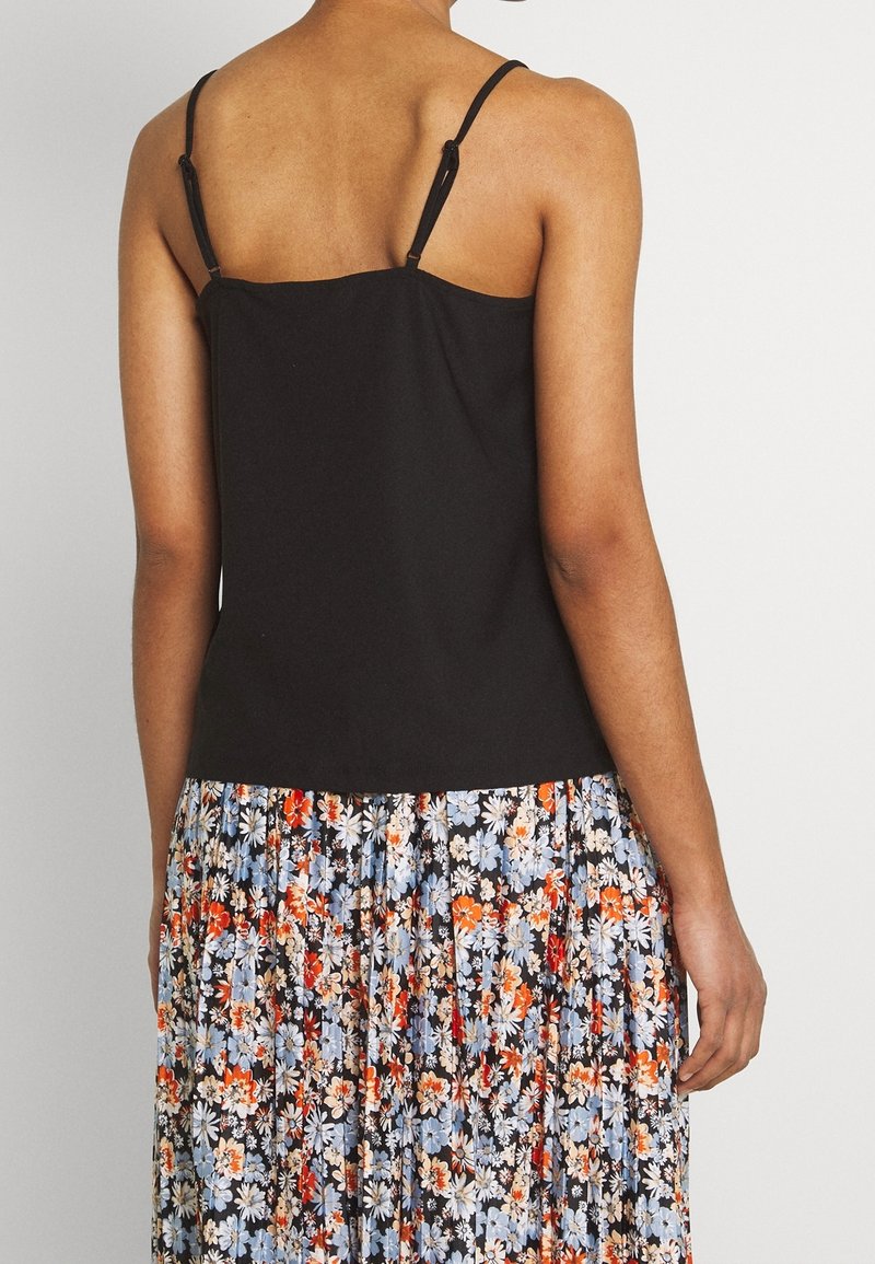 Femme portant un haut noir sans manches et une jupe plissée fleurie avec des fleurs bleues, orange et blanches, debout, de dos sur fond blanc.