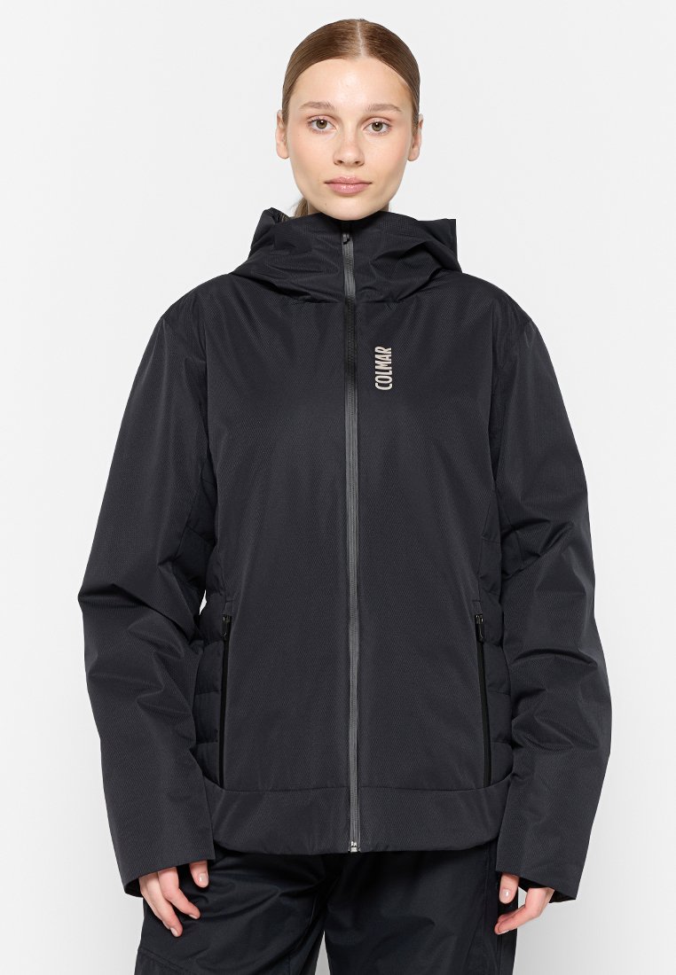 Donna che indossa un giubbotto con zip nera e cappuccio con il logo Colmar sul petto, in piedi contro uno sfondo bianco semplice.