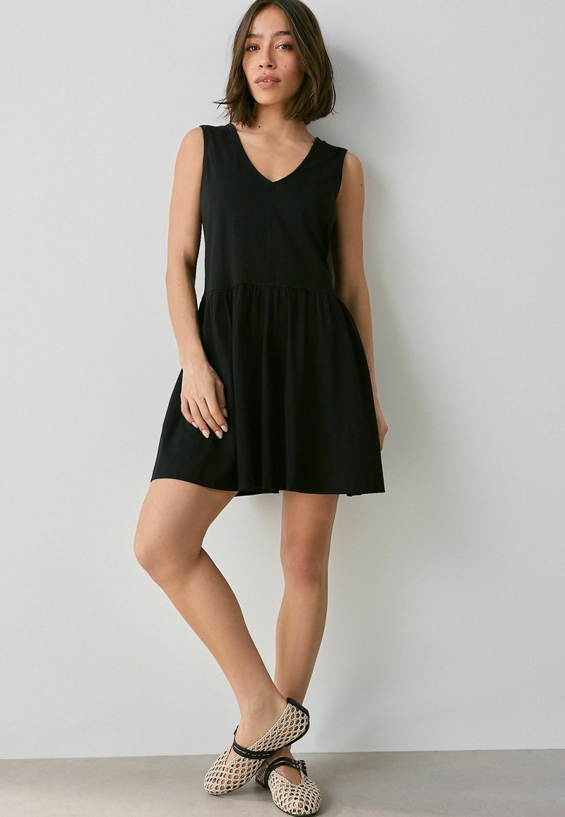 Schwarzes ärmelloses Kleid mit V-Ausschnitt, geraffter Taille und schwingendem Rock, getragen mit beigen, gemusterten Schuhen mit schwarzen Akzenten.