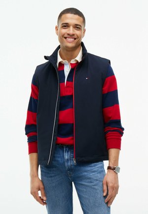 Ung mand smilende, iført en marineblå vest over en rød og marineblå stribet langærmet skjorte med blå jeans og et armbåndsur.