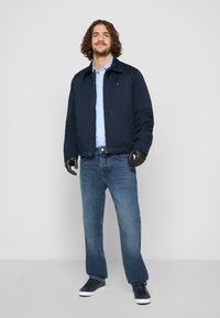 Marine bomberjakke, lyseblå skjorte under, mørkeblå jeans og svarte hansker. Svarte joggesko med hvite såler. Minimalistisk design.