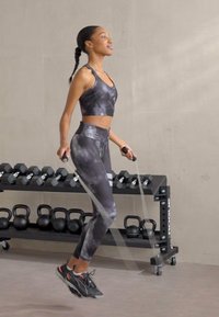 Sportovní podprsenka a legíny s černým mramorovým vzorem; uživatel skáče přes švihadlo a má na sobě černé atletické boty; v pozadí je viditelné gym vybavení.