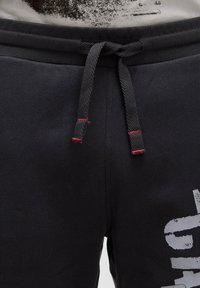 Schwarze Trainingshose mit einem Kordelzugbund, versehen mit roten Akzenten und einem großen weißen Grafikdruck am linken Bein. Glattes Baumwollgewebe.