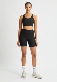 Un reggiseno sportivo nero con costine e pantaloni corti a vita alta, entrambi aderenti e elasticizzati. La modella indossa scarpe da ginnastica grigie e calzini bianchi alla caviglia.