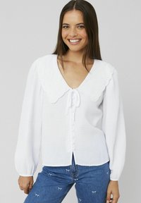 Blouse blanche avec un grand col festonné, en tissu texturé, à manches longues, et fermeture à boutons sur le devant, associée à un jean en denim bleu.