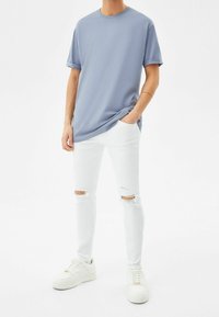 Camiseta de algodón azul claro con mangas enrolladas, combinada con jeans ajustados blancos con rodillas desgastadas y zapatillas blancas, sobre un fondo neutro.