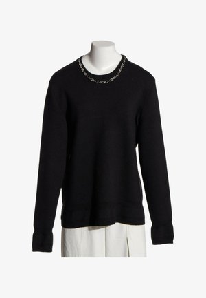 Pull noir à manches longues avec un collier chaîne argentée sur un torse de mannequin blanc, associé à un pantalon plissé blanc.