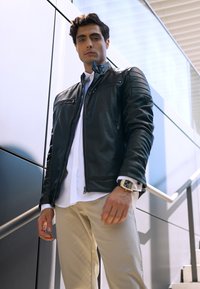Juodas odinis švarkas su vertikaliais siūlėmis ant pečių, užtrauktukais ant kišenių ir sidabriniais detalėmis, derinamas su balta marškiniais ir smėlio spalvos kelnėmis.