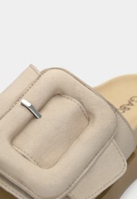 Παντόφλα μπεζ με απλή εφαρμογή, με απαλό επάνω μέρος από σουέντ, έντονο τετράγωνο αγκράφα και υφασμένο πέλμα με λεπτομέρειες ραφής.