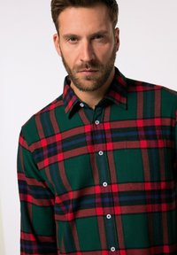 Grøn flannel skjorte med rød, sort og marineblå tern. Har knaplukning, spidse krave og en patchlomme på brystet.