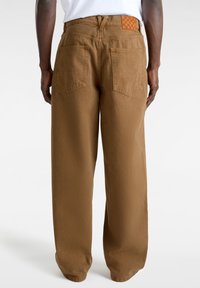 Pantalones de pierna ancha en marrón, hechos de suave algodón, con dos bolsillos traseros y un distintivo parche de tela a cuadros en color beige en la cinturilla.
