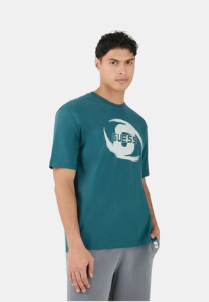 Giovane uomo con capelli corti e ricci che indossa una t-shirt color verde acqua con il logo GUESS e pantaloni grigio chiaro, in piedi davanti a uno sfondo bianco semplice.