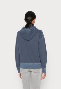 Blå hoodie med en stor huva, ribbad nederkant och muddar. Mjuk tyg med en något blekt textur, som har kontrasterande sömmar.