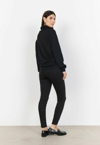 Haut à col roulé noir et jean skinny noir ; le modèle se tient de profil, montrant le tissu lisse et la silhouette ajustée, associé à des mocassins brillants.