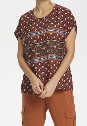 Femme portant un chemisier rouge à manches courtes avec des motifs géométriques et floraux, associé à un pantalon cargo couleur rouille.