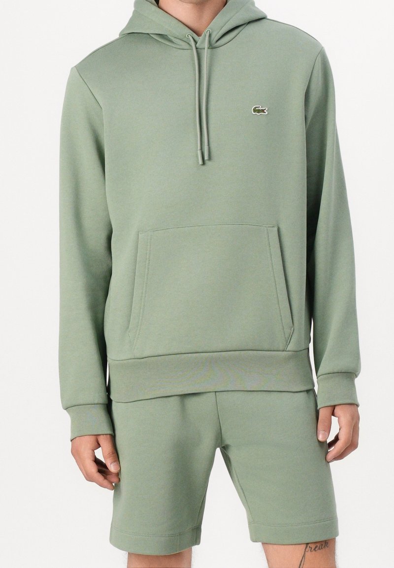 Homme portant un sweat à capuche vert clair avec poche avant et short assorti, tatouage visible sur le bas de la jambe.