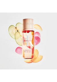 Flacon de parfum en verre transparent, liquide couleur pêche, avec un bouchon beige. Entouré de pêches tranchées et d'une pomme verte, sur un fond blanc.
