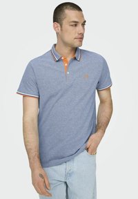 Polo de color azul claro con detalles en naranja en el cuello y las mangas, que cuenta con una tapeta de tres botones y un pequeño logo en el pecho.