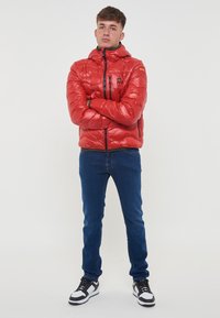 Giacca a vento rossa con finitura lucida, chiusura frontale con zip e cappuccio; presenta dettagli neri sulle zip e una tasca sul petto. Indossata con jeans blu e scarpe da ginnastica.
