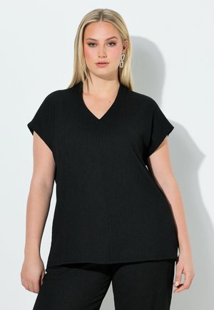 Blonde vrouw in zwarte top met V-hals en broek staat tegen een witte muur, draagt rechthoekige oorbellen met schakels.